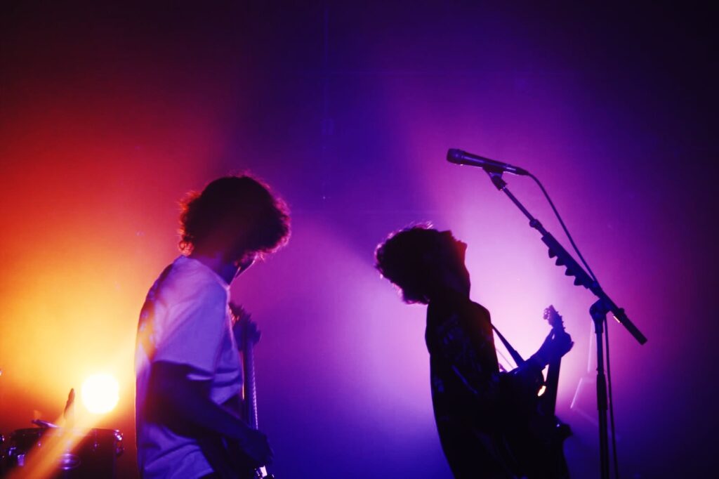 Deux membres du groupe GEHEN en silhouette sur scène pendant un concert, éclairés par des lumières colorées.